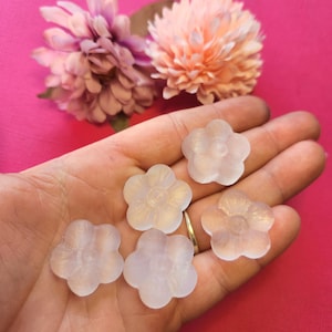 May include: A hand displays six translucent, flower-shaped beads. The beads are a soft, pearlescent white with subtle details. Two artificial flowers in shades of pink and peach are in the background against a bright pink backdrop.