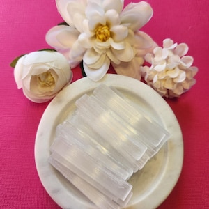 May include: A display of clear selenite sticks in a white marble dish, with white artificial flowers set against a vibrant pink backdrop. The selenite crystals are rectangular and translucent.