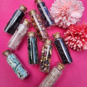 May include: An assortment of small glass bottles, each containing different coloured gemstones. The bottles have cork stoppers and are arranged on a pink surface. The gemstones include black, white, blue, and brown stones. Pink and red flowers are in the background.