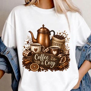 Peut inclure: Un t-shirt blanc avec une impression graphique représentant une cafetière en cuivre, deux tasses à motifs de cœur, un sac en toile de jute marqué Coffee, des brins de coton, des bâtons de cannelle et des tranches d'orange. Le texte Coffee & Cozy est centré sur un panneau en bois.