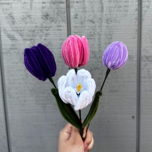 May include: A bouquet of handmade pipe cleaner tulips in various colors. The flowers include a deep purple, bright pink, light purple, and white with a yellow center. The stems are green, and the flowers are held together.