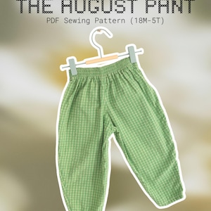 May include: A pair of green and white checkered pants with an elastic waistband, hanging on a light-colored hanger. The text above reads THE AUGUST PANT PDF Sewing Pattern (18M-5T) in a cross-stitch font style against a blurred background.