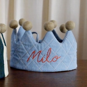 May include: Two fabric crowns. One is striped in navy and white with the name "August" embroidered in gold. The other is light blue with a quilted pattern and the name "Milo" embroidered in red. Each crown has beige pom-poms.