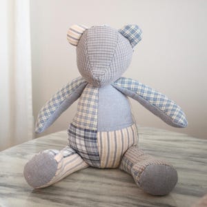 May include: A patchwork teddy bear in blue, grey, and cream. The bear is made from various patterned fabrics, including plaid and stripes. It has a soft appearance and is sitting on a marble surface. The bear is a charming handmade item.