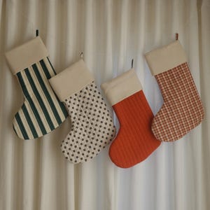 Op de afbeelding: Vier kerstsokken hangen voor een wit gordijn. De sokken hebben verschillende patronen: groen-witte strepen, kleine zwarte stippen op wit, gewatteerd oranje en rood-wit geruit. Elke sok heeft een crèmekleurige manchet.