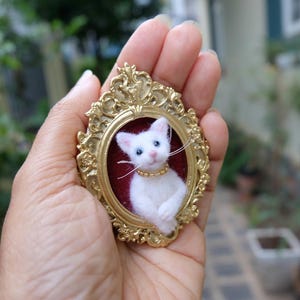 May include: A small, oval-shaped gold-colored frame with ornate detailing, featuring a miniature white felted cat figure inside. The cat has blue eyes, a pink nose, and a gold-colored beaded collar, set against a deep red velvet-like background.