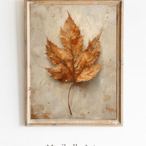 Puede incluir: Una impresión enmarcada que presenta una representación detallada y texturizada de una gran hoja de otoño marrón. La hoja está centrada contra un fondo neutro y texturizado, dentro de un marco de madera de tono claro. El texto "Maribelle Arts Printable Arts" está en la parte inferior.