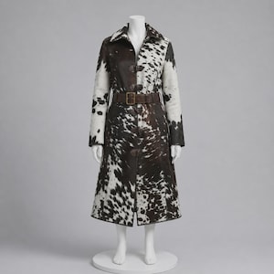 May include: A long coat featuring a brown and white cowhide print pattern. The garment includes a pointed collar, long sleeves, and a dark brown leather belt with a rectangular buckle at the waist. The front closure is secured with toggle fasteners.
