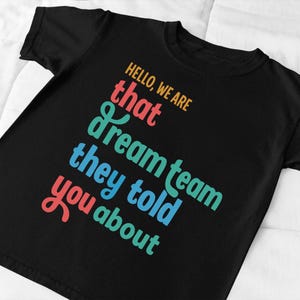 May include: Black t-shirt with the text "HELLO, WE ARE that dream team they told you about" in orange, red, teal, and blue. The text is arranged diagonally across the shirt.