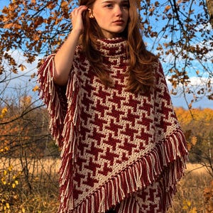 May include: A crochet poncho featuring a two-tone houndstooth pattern in deep red and cream. The garment has a high turtleneck collar and long, thick fringe detailing along the edges, creating a textured, layered appearance.