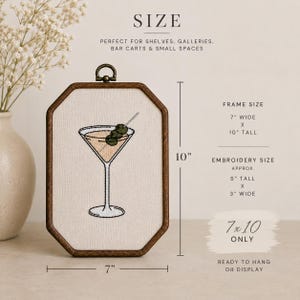 May include: An octagonal wooden frame containing embroidery of a martini glass with two green olives on a pick. The frame measures 7 inches wide by 10 inches tall, while the embroidery measures approximately 5 inches tall by 3 inches wide.