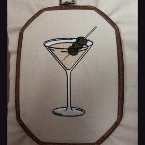 May include: An embroidery hoop art piece featuring a stitched martini glass filled with a light tan liquid and garnished with three dark green olives on a silver pick. The design is centered on off-white fabric within a dark brown wooden frame.