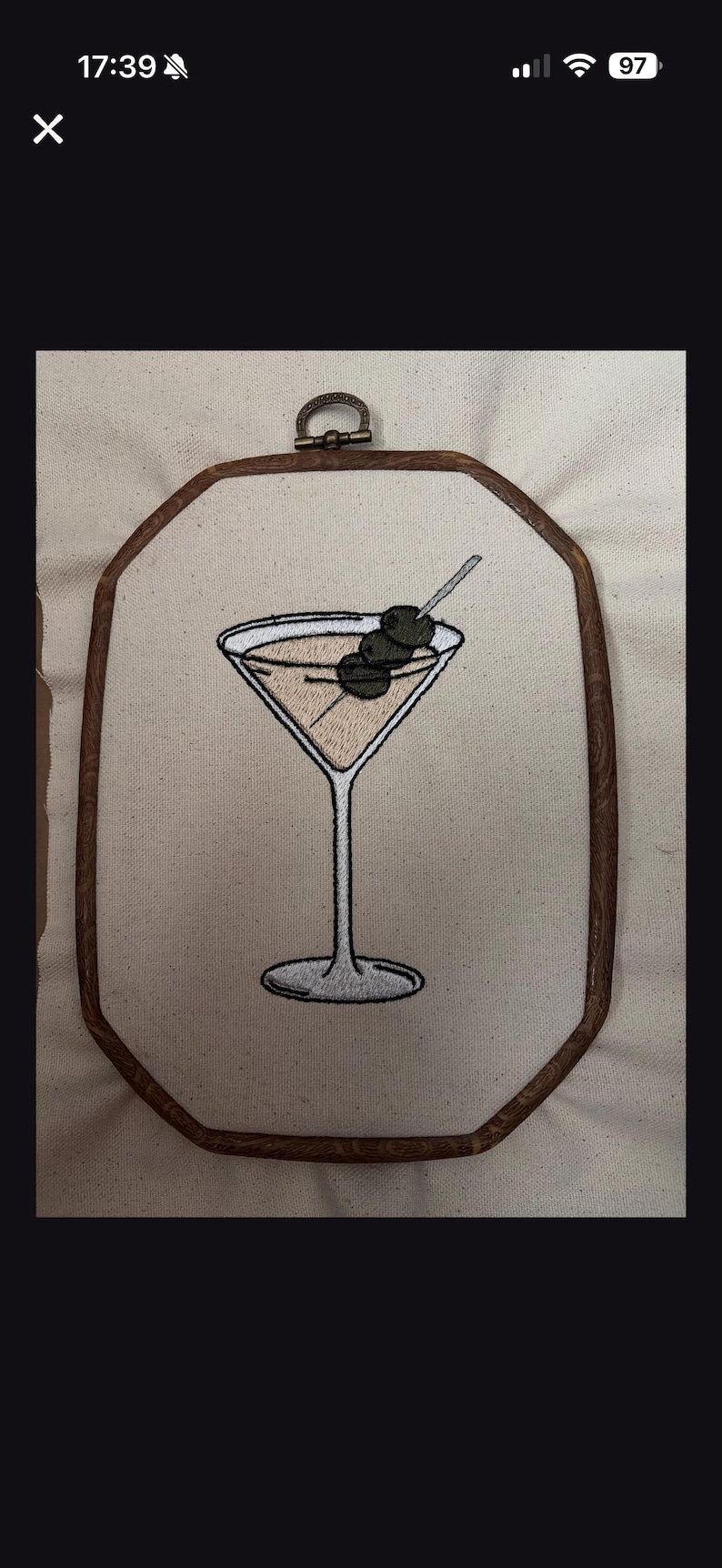 May include: An embroidery hoop art piece featuring a stitched martini glass filled with a light tan liquid and garnished with three dark green olives on a silver pick. The design is centered on off-white fabric within a dark brown wooden frame.