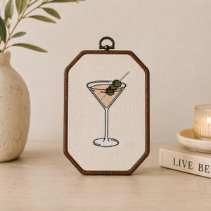 May include: An octagonal wooden embroidery hoop frame containing a fabric panel with a stitched martini glass design. The cocktail is light tan with three green olives on a silver pick. The frame features a small metal hanging loop at the top.