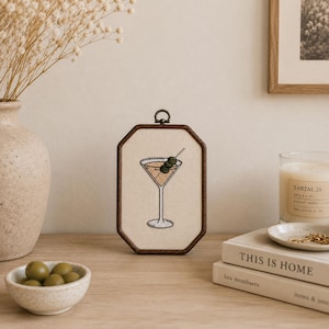 May include: An octagonal wooden embroidery hoop frame containing a cream-colored fabric panel with a stitched illustration of a martini glass filled with a light brown liquid and garnished with three green olives on a silver cocktail pick.