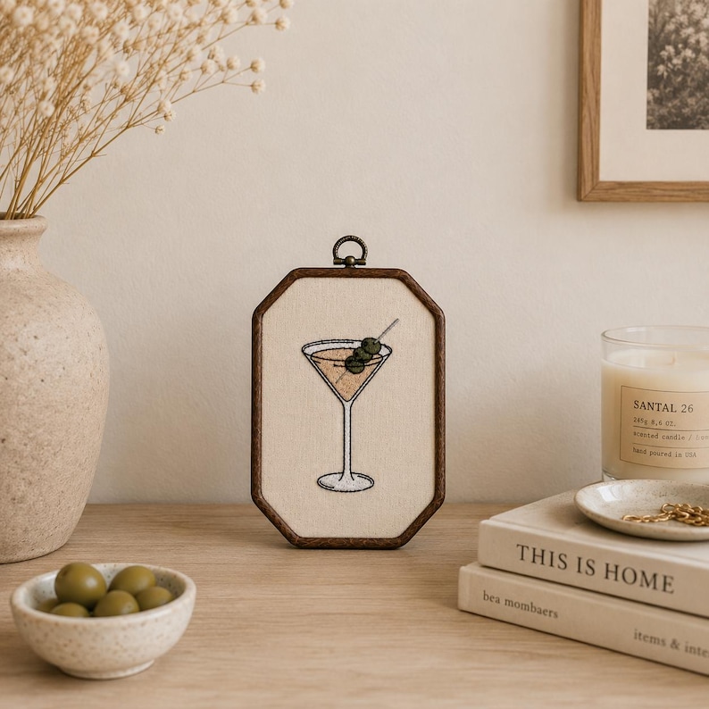 May include: An octagonal wooden embroidery hoop frame containing a cream-colored fabric panel with a stitched illustration of a martini glass filled with a light brown liquid and garnished with three green olives on a silver cocktail pick.