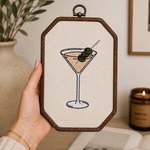 May include: An embroidery hoop art piece featuring a stitched martini glass filled with a light tan liquid and garnished with three green olives on a silver toothpick. The design is centered on off-white fabric within a dark brown wooden frame.
