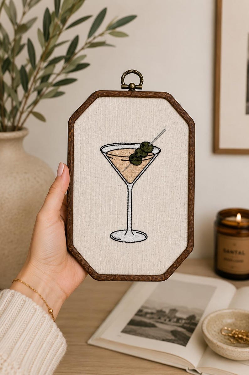 May include: An embroidery hoop art piece featuring a stitched martini glass filled with a light tan liquid and garnished with three green olives on a silver toothpick. The design is centered on off-white fabric within a dark brown wooden frame.