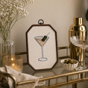 May include: An octagonal wooden frame containing an embroidered martini glass with three green olives on a toothpick. The embroidery features a tan liquid, a white glass outline, and dark green olives centered on a cream-colored fabric background.