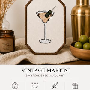 May include: An octagonal wooden embroidery hoop containing a cream-colored fabric panel with a hand-stitched martini glass design featuring three green olives on a pick. The text VINTAGE MARTINI EMBROIDERED WALL ART is printed below the hoop.