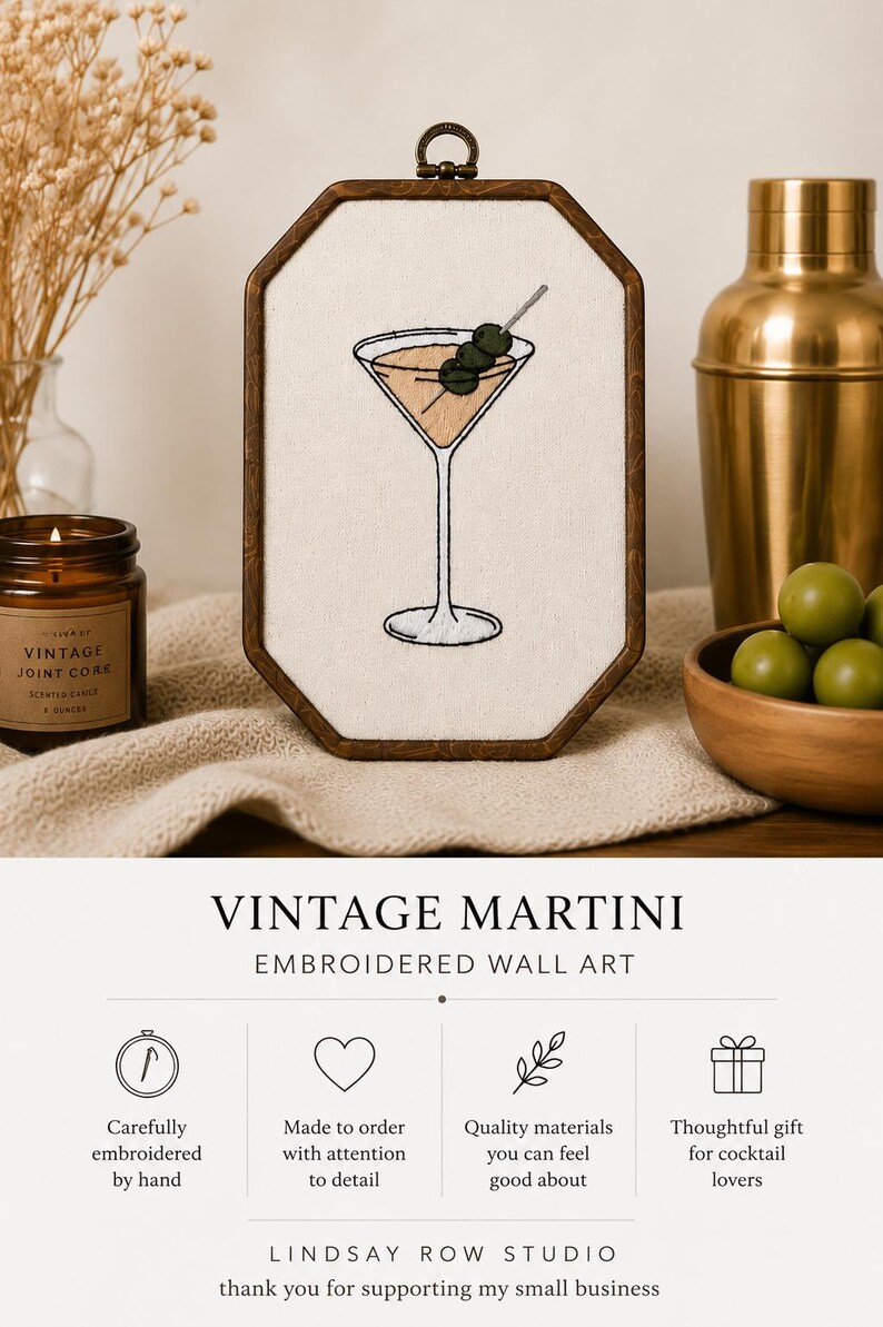 May include: An octagonal wooden embroidery hoop containing a cream-colored fabric panel with a hand-stitched martini glass design featuring three green olives on a pick. The text VINTAGE MARTINI EMBROIDERED WALL ART is printed below the hoop.