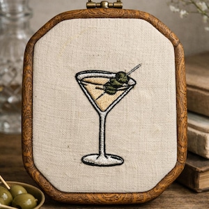 May include: An embroidery hoop featuring a stitched martini glass filled with a pale yellow liquid and garnished with two green olives on a toothpick. The design is centered on off-white fabric and framed by a dark brown wooden octagonal hoop.