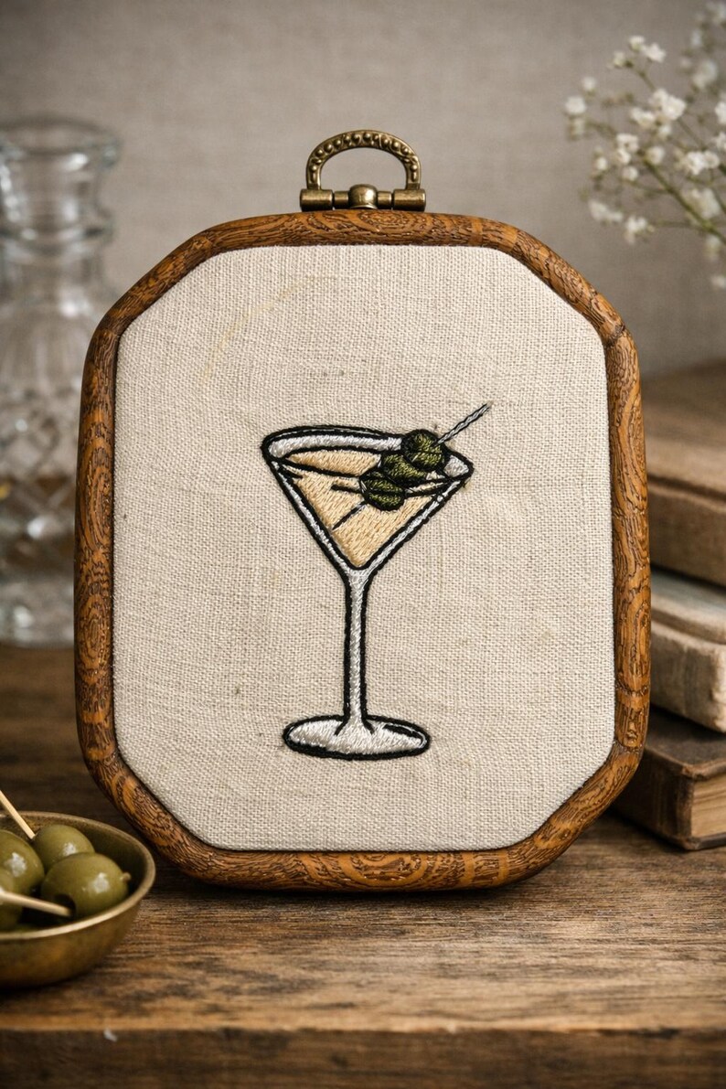 May include: An embroidery hoop featuring a stitched martini glass filled with a pale yellow liquid and garnished with two green olives on a toothpick. The design is centered on off-white fabric and framed by a dark brown wooden octagonal hoop.