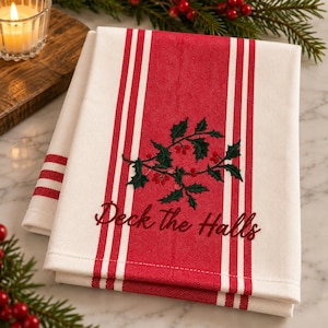 May include: A folded white kitchen towel featuring a wide red vertical stripe down the center with thin red stripes on either side. The towel is embroidered with a green holly branch with red berries and the text Deck the Halls in red script.