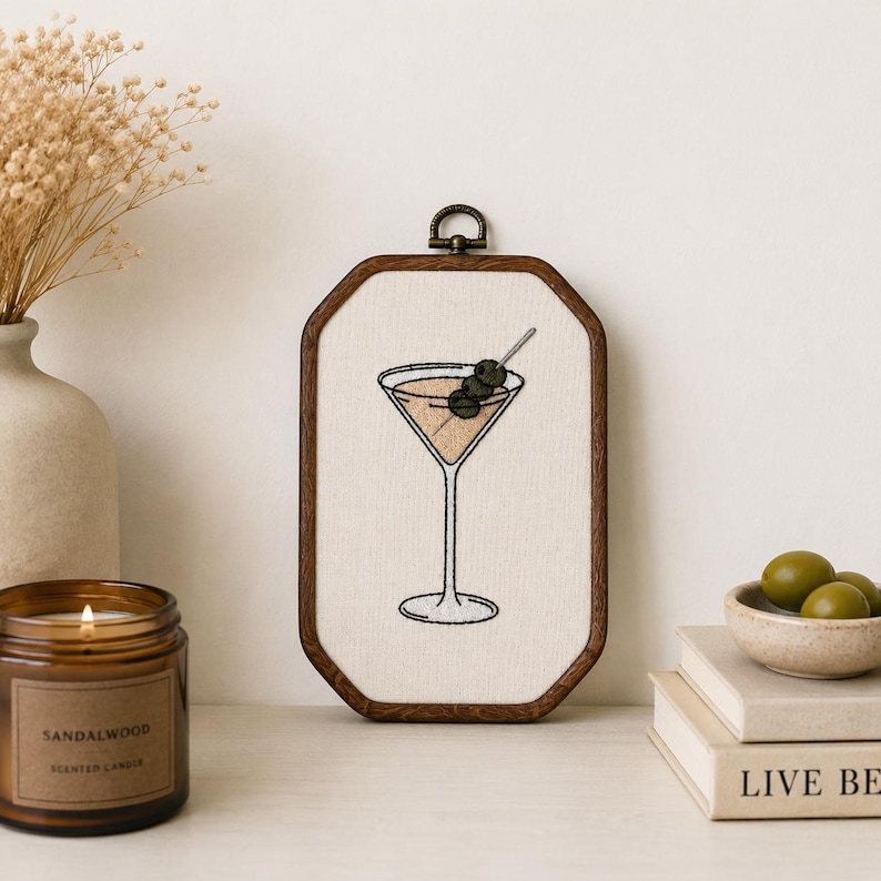 May include: An octagonal wooden embroidery hoop frame containing a cream-colored fabric panel with a hand-stitched martini glass design. The cocktail is depicted with a light tan liquid and two dark green olives on a silver toothpick skewer.