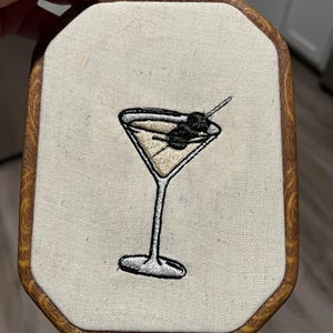 May include: An embroidery hoop featuring a stitched martini glass with a light tan liquid and a dark olive on a toothpick. The fabric is off-white, and the frame is an octagonal wooden hoop with a textured, light brown wood grain pattern.