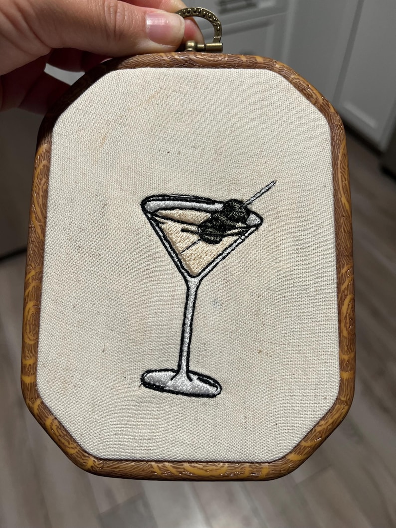 May include: An embroidery hoop featuring a stitched martini glass with a light tan liquid and a dark olive on a toothpick. The fabric is off-white, and the frame is an octagonal wooden hoop with a textured, light brown wood grain pattern.