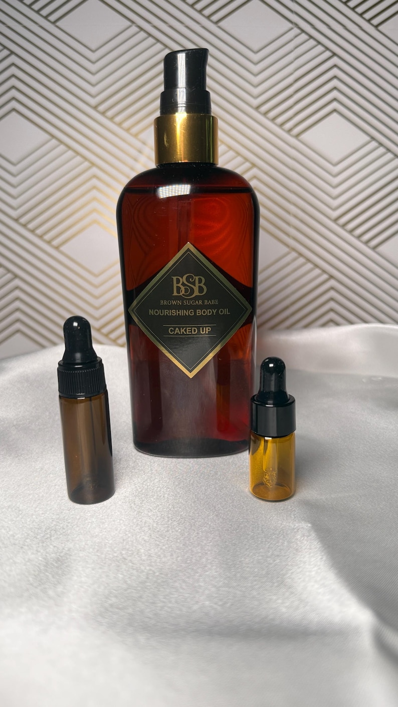 May include: A brown body oil bottle with a gold cap and a black label that reads 'Brown Sugar Babe Nourishing Body Oil Caked Up'. Two smaller amber glass bottles with black droppers are also present. The background is a white surface with a gold and white geometric pattern.