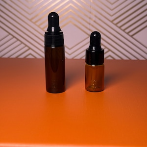 May include: Two amber glass dropper bottles with black caps. The bottles are different sizes, with one taller than the other. They are set against an orange surface with a pink and gold geometric patterned background.