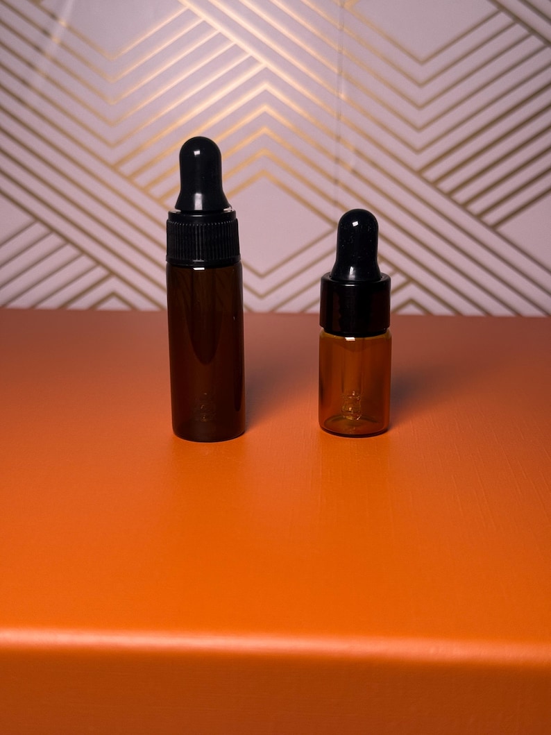 May include: Two amber glass dropper bottles with black caps. The bottles are different sizes, with one taller than the other. They are set against an orange surface with a pink and gold geometric patterned background.