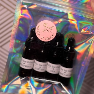 May include: A clear, iridescent bag containing four small, dark glass bottles with black droppers. A pink sticker with gold text that says "Thank you" is on the bag. The bottles have white labels with black text.
