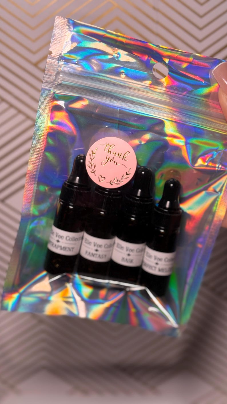 May include: A clear, iridescent pouch containing four small, dark glass bottles with black droppers. A pink sticker with gold text that says "Thank you" is on top. The bottles have white labels with text.