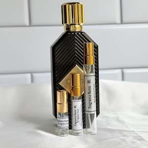 May include: A large black glass perfume bottle with a gold cap and a chevron pattern, accompanied by three smaller clear glass spray vials with gold caps and white labels reading Fragrance Name - ML by Fragrance House.