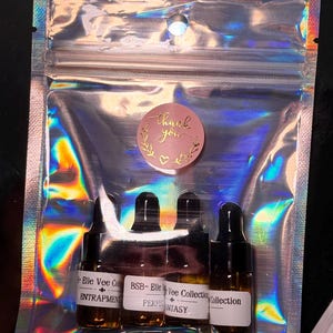 May include: A clear, iridescent pouch containing four small, dark glass bottles with black caps. Each bottle has a white label with text. A pink sticker with the words "thank you" is on the pouch.