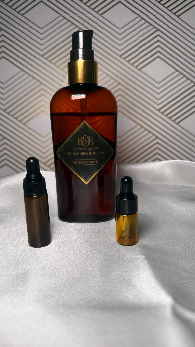 May include: A brown glass bottle of body oil with a black and gold pump, flanked by two smaller dropper bottles. The label reads "Brown Sugar Babe Nourishing Body Oil" and "MS Independent". The background is a white surface with a gold geometric pattern.