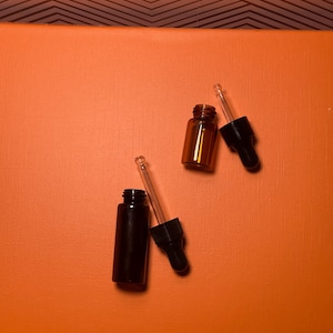 May include: Two small amber glass bottles and one black glass bottle with black droppers. The bottles are on an orange surface. The background is a geometric pattern in brown and white.