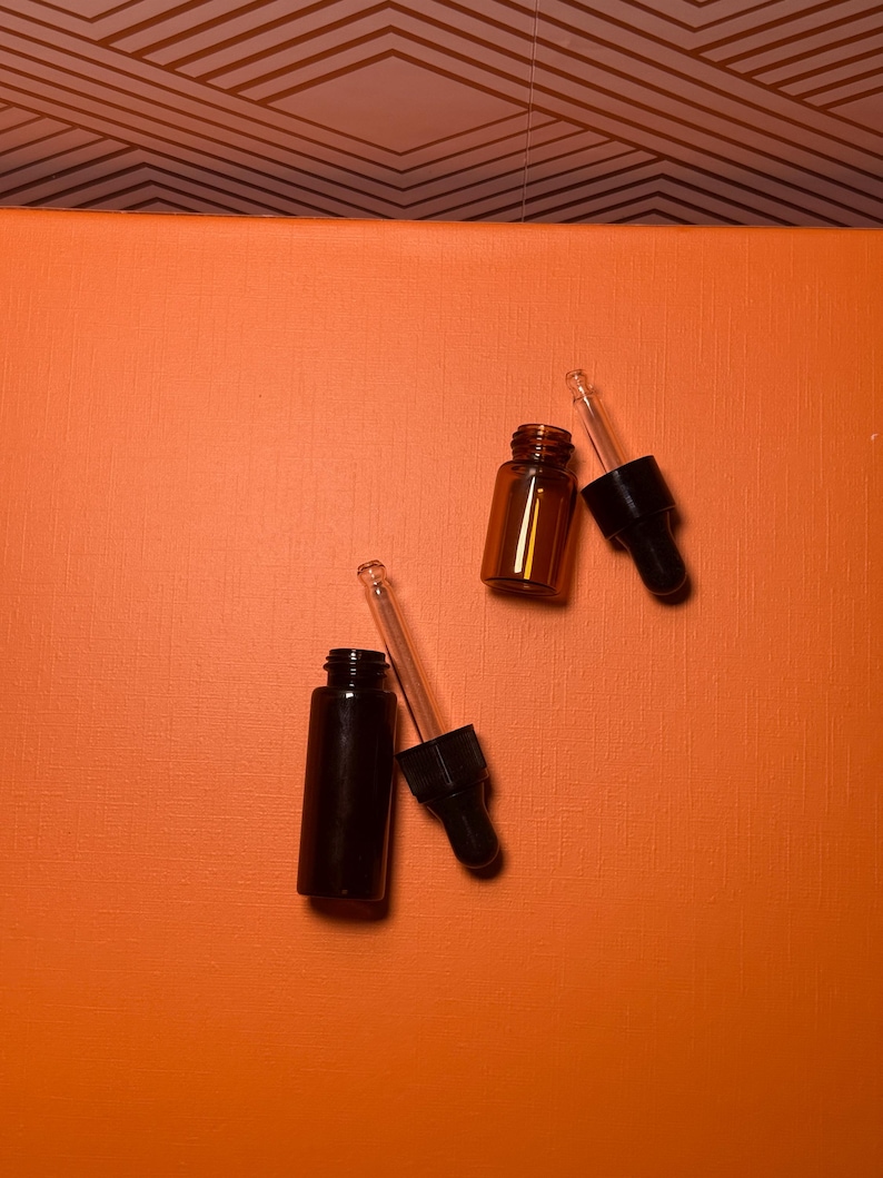 May include: Two small amber glass bottles and one black glass bottle with black droppers. The bottles are on an orange surface. The background is a geometric pattern in brown and white.