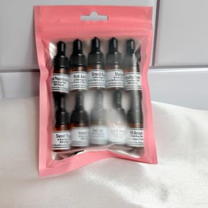May include: A pink plastic resealable pouch containing ten small amber glass vials with black dropper caps. Each vial features a white label with black text identifying various fragrance names including Pink Poundcake, Rich Aura, and Mi Amore.