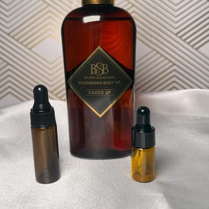 May include: A brown glass bottle of Brown Sugar Babe Nourishing Body Oil with a black and gold label, and two smaller dropper bottles. The label reads "CAKED UP". The bottles are on a white surface.