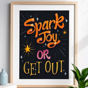 以下が含まれることがあります： 黒い背景に、オレンジ色、ピンク色、黄色の文字で「Spark Joy OR GET OUT」と書かれた額入りのアートプリント。 プリントは明るい木製のフレームに入っており、小さな青い花瓶と植物が置かれた木製の棚に飾られています。