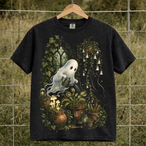 May include: A black short-sleeve t-shirt featuring a graphic print of a white ghost watering a Venus flytrap with a small watering can. The scene includes potted plants, glowing mushrooms, hanging bell-shaped flowers, and a black cat silhouette.