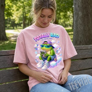 May include: A light pink t-shirt featuring a graphic of a green frog wearing a purple baseball cap and blue sunglasses. The frog holds a handheld gaming console, sits on a cloud, and is surrounded by CDs, stars, and the text TOTALLY RAD (at being sad).