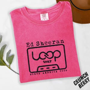 May include: A pink Comfort Colors t-shirt featuring a black graphic of a cassette tape with the words "Ed Sheeran Loop Tour North America 2026." The shirt has a crew neck and short sleeves. A brown hat is visible in the background.
