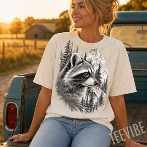 May include: A cream-colored short-sleeve t-shirt featuring a detailed black and white graphic print of a raccoon surrounded by pine trees and mountain peaks. The design is centered on the chest, showcasing the animal in profile with textured fur.