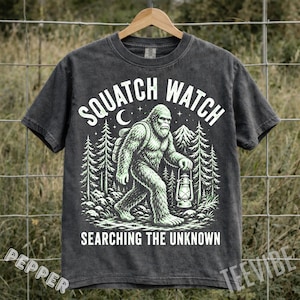 May include: Dark gray t-shirt with a graphic of a Bigfoot carrying a lantern and backpack, with the words "SQUATCH WATCH" above and "SEARCHING THE UNKNOWN" below. The design is in a light green color.