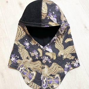 May include: A hooded neck warmer featuring a black denim panel on the crown and a dark fabric body printed with gold eagles, purple cherry blossoms, and geometric patterns. The front closure includes three metal snap buttons and a black lining.